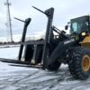 Loader forklift lifting large black metal beams in snowy area.