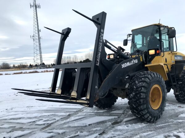 Loader forklift lifting large black metal beams in snowy area.