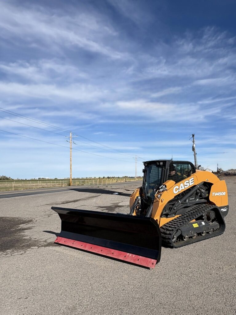 A tracked snowplow vehicle parked on a paved road under a blue sky.